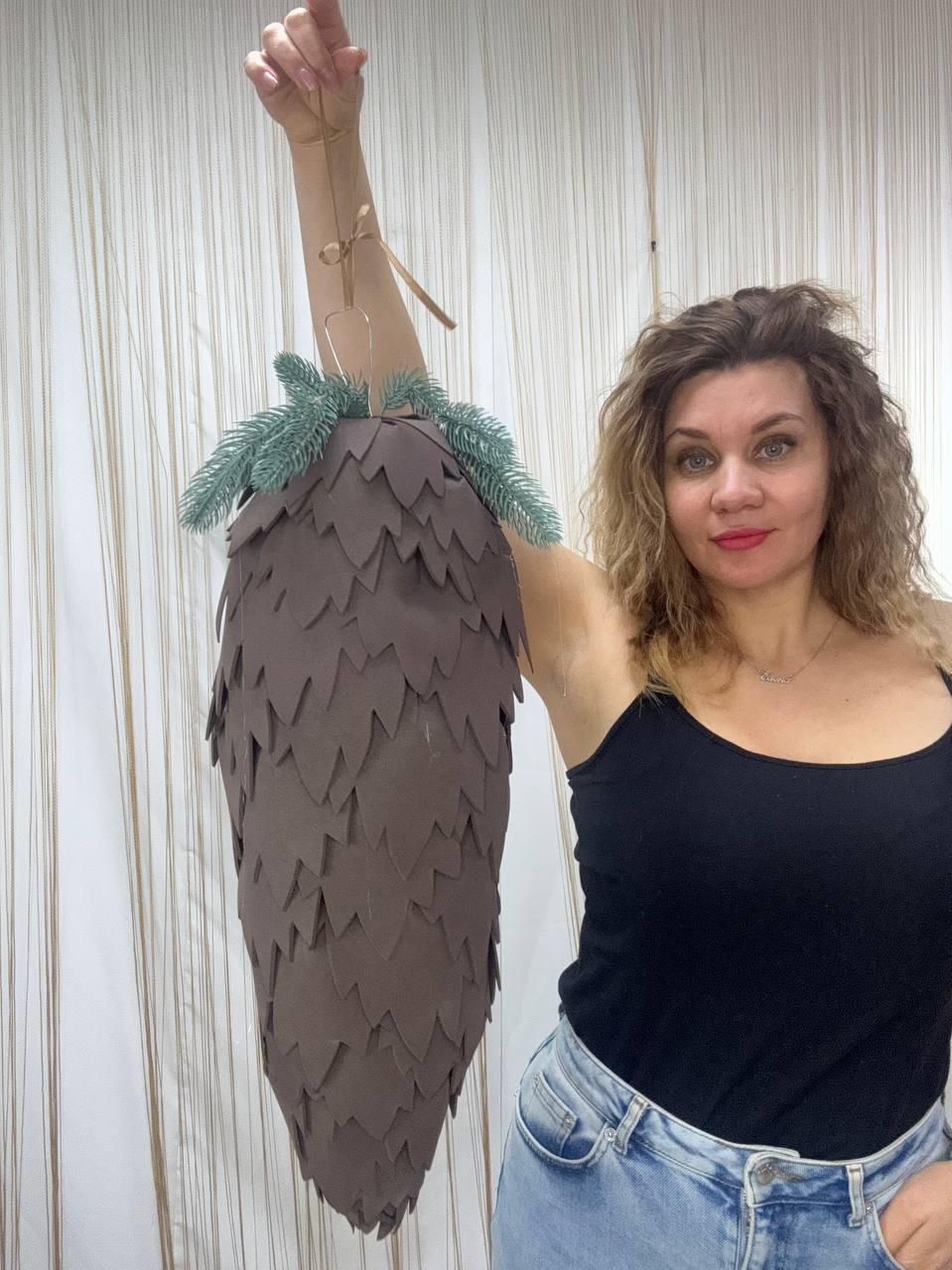 May include: A large, brown, cone-shaped ornament with a textured, layered design. The top is adorned with green faux pine branches. The ornament is suspended by a thin, natural-colored string, suggesting it's for hanging.