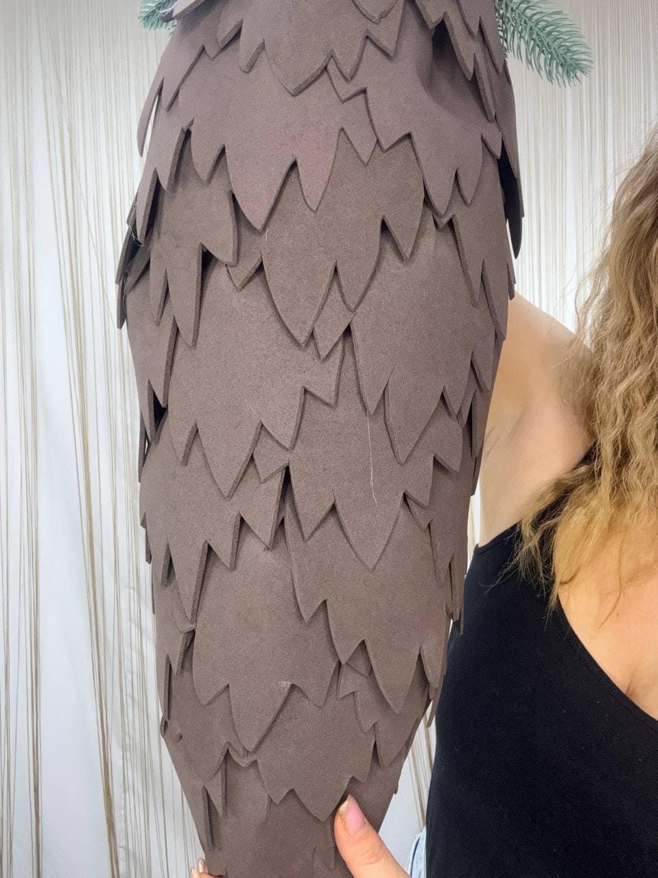 May include: A large, brown, cone-shaped decoration with a layered, scale-like design. The decoration has a pointed top and a rounded base. A small sprig of green and blue faux foliage is attached to the top. The decoration is held by a person.