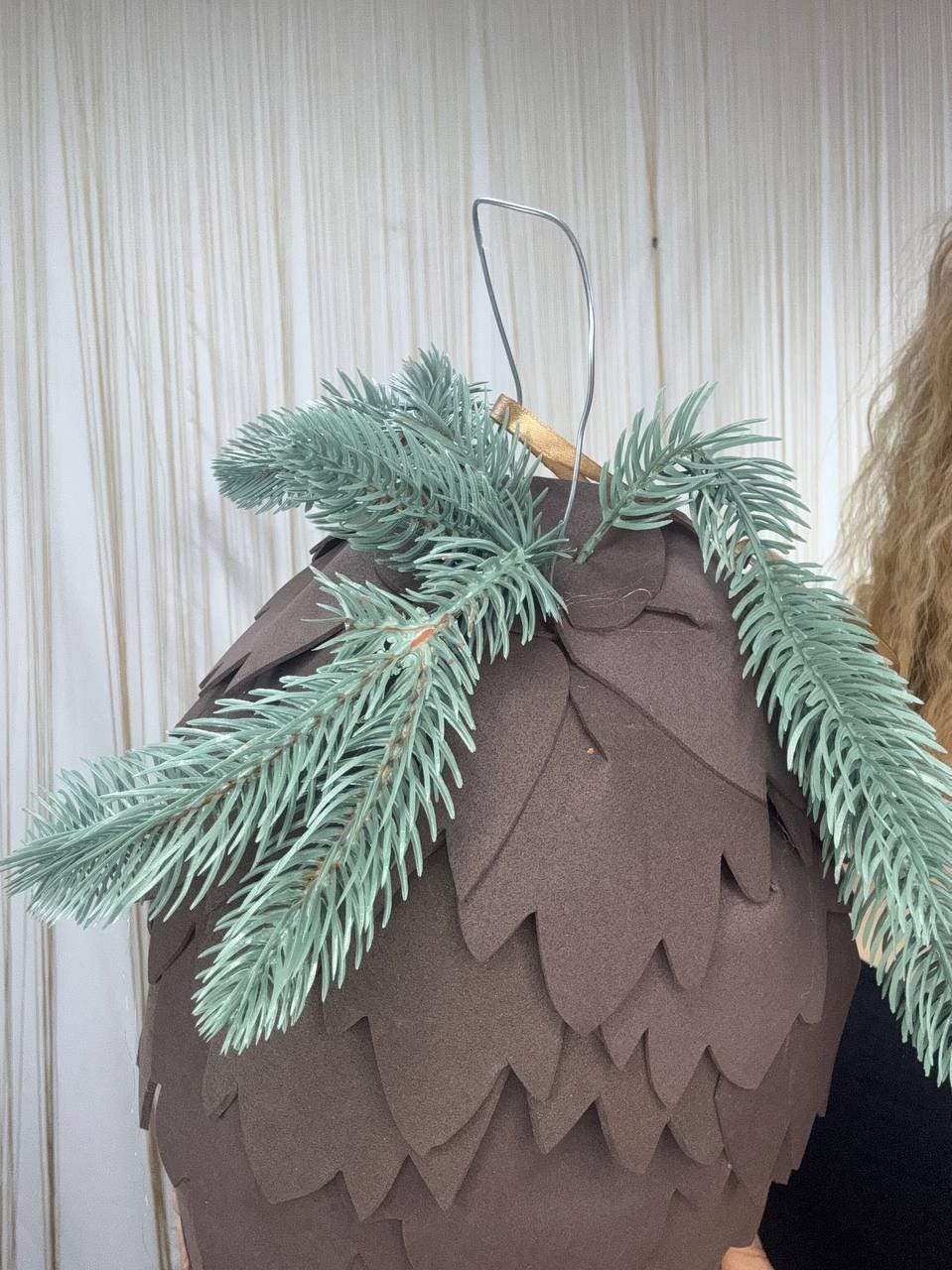 May include: A brown, cone-shaped ornament with a metal hanger and faux evergreen branches. The ornament is made of layered, petal-shaped pieces. The evergreen branches are a muted green color.