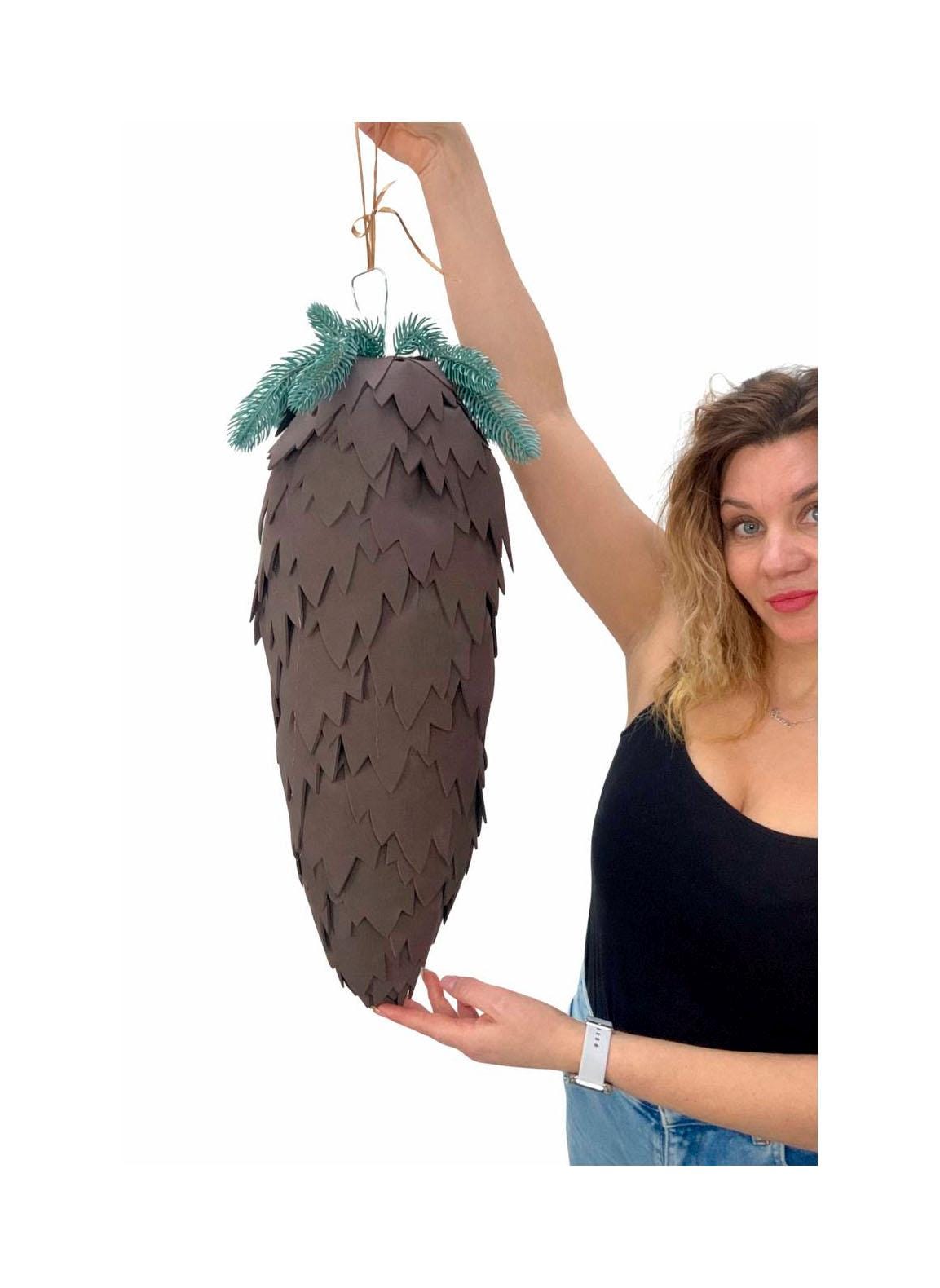 May include: A large, brown paper pinecone decoration with a textured surface. The pinecone is topped with green faux pine needles and suspended by a thin, tan string. A unique, handcrafted holiday or seasonal decoration.