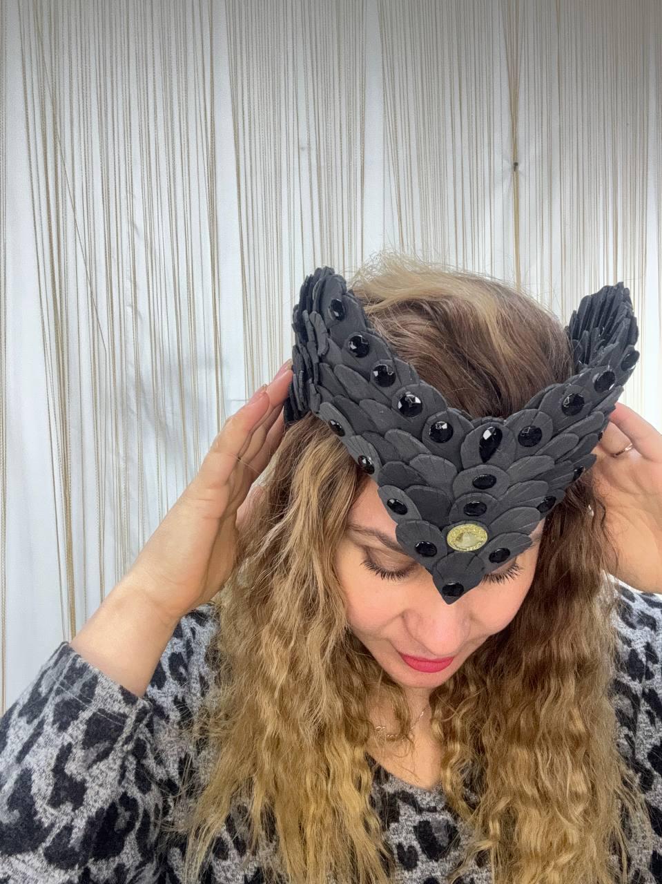 May include: A black headpiece with a layered, scale-like design and embellished with black gemstones. The headpiece has a central medallion and is being adjusted by a person.