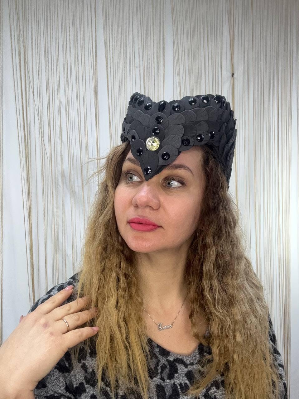 May include: A black headpiece with a leaf-like design, adorned with black beads and a central gold-colored gem. The headpiece is worn by a person with long, wavy blonde hair and a patterned top.