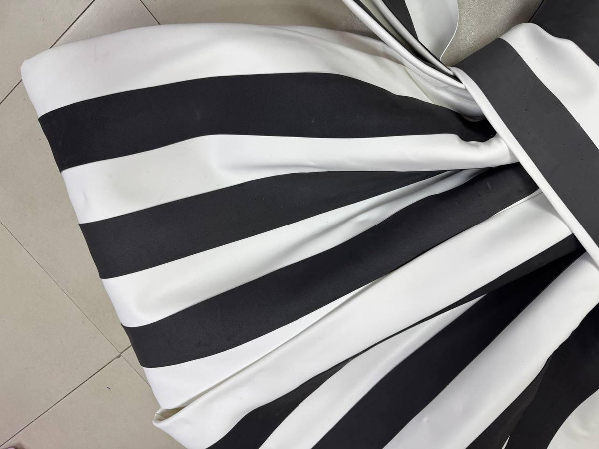 May include: Close-up of a fabric with bold black and white stripes. The fabric is folded and gathered, creating a textured effect. The stripes are evenly spaced and run across the material, creating a striking visual pattern. The fabric appears to be smooth and possibly satin.