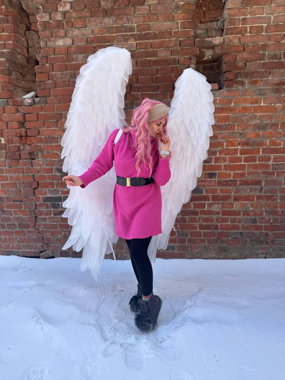 May include: A person wearing a pink dress with a black belt and black leggings, posing in front of a brick wall. Large white angel wings are attached to the person's back. The person is standing in the snow.