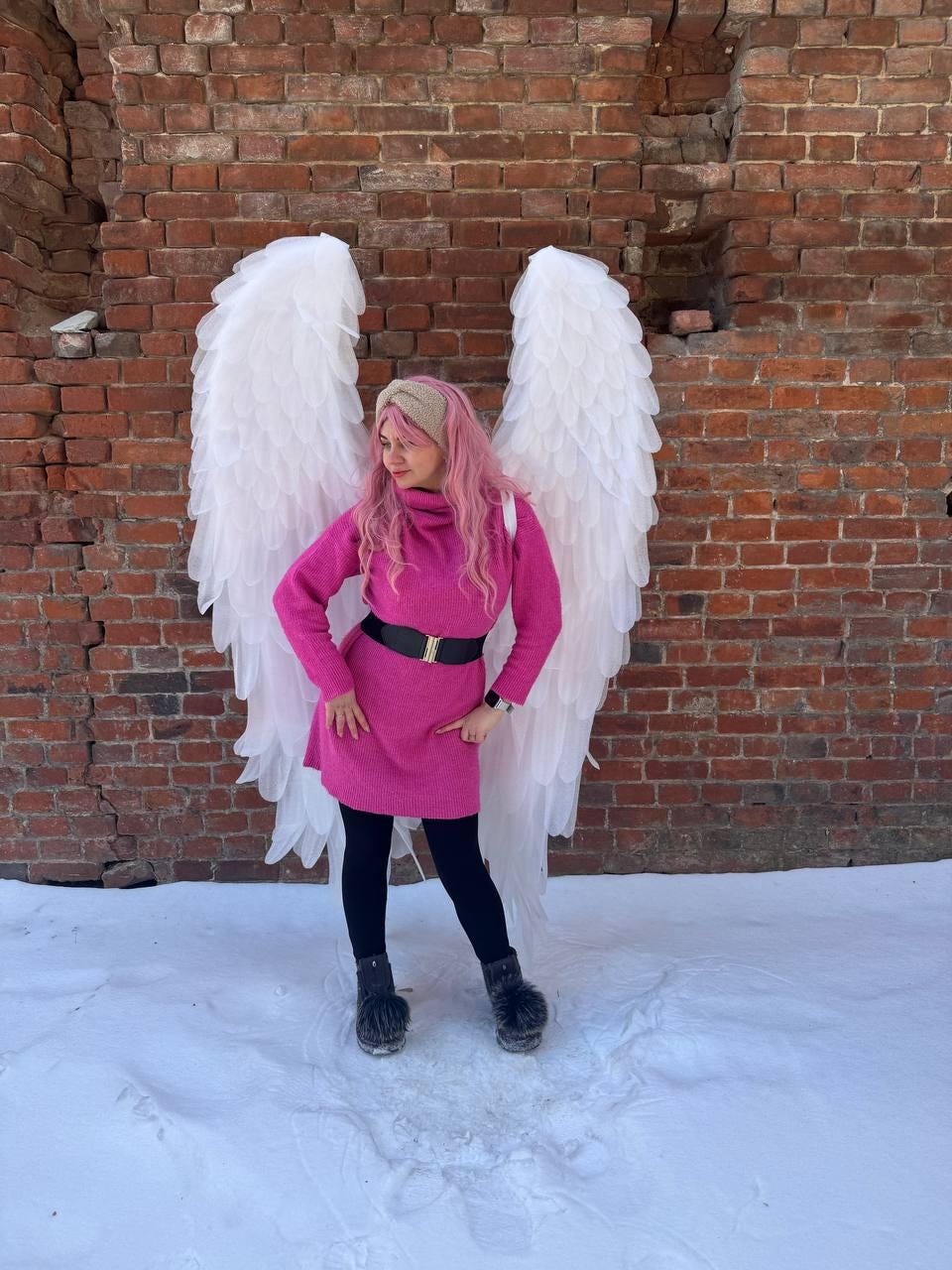 May include: A person wearing a pink dress with a black belt, black leggings, and furry boots stands in front of large white angel wings. The background is a weathered brick wall and snow.