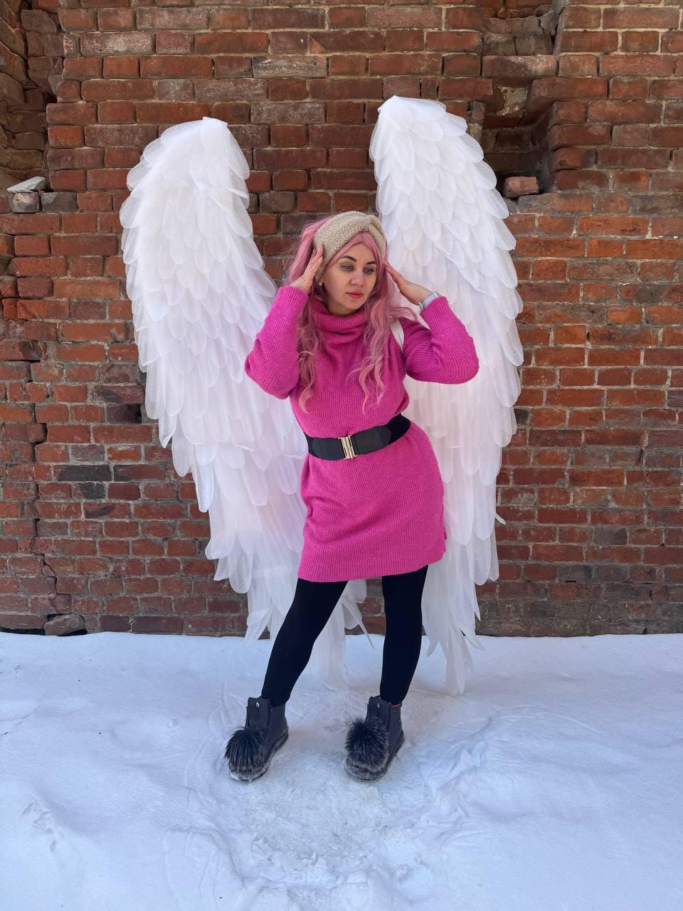 May include: A person wearing a pink turtleneck sweater dress with a black belt and black leggings, standing in front of large white angel wings. The background is a brick wall and snow.