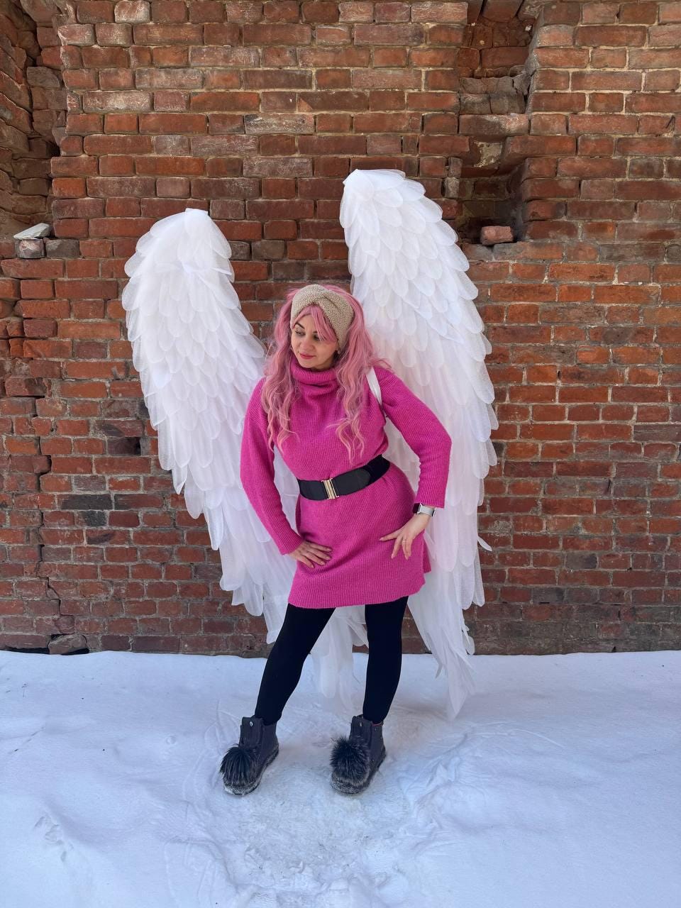 May include: A person wearing a pink sweater dress, black leggings, and black boots with white angel wings against a brick wall. The person has pink hair and a beige headband. A black belt cinches the waist.