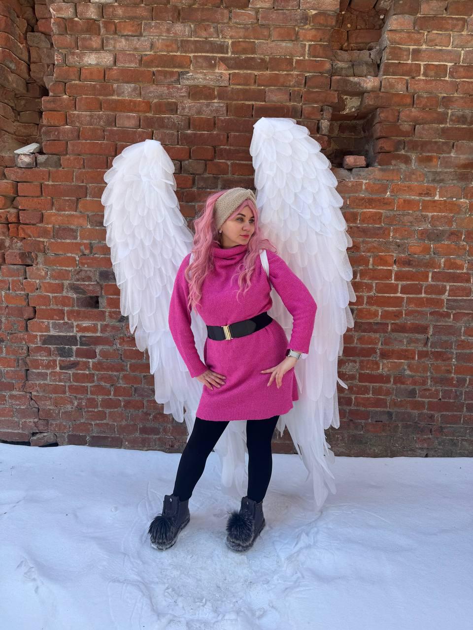 May include: A person wearing a pink sweater dress, black leggings, and black boots with fur trim, posing in front of a brick wall. Large white angel wings are behind the person. A black belt cinches the waist of the dress.