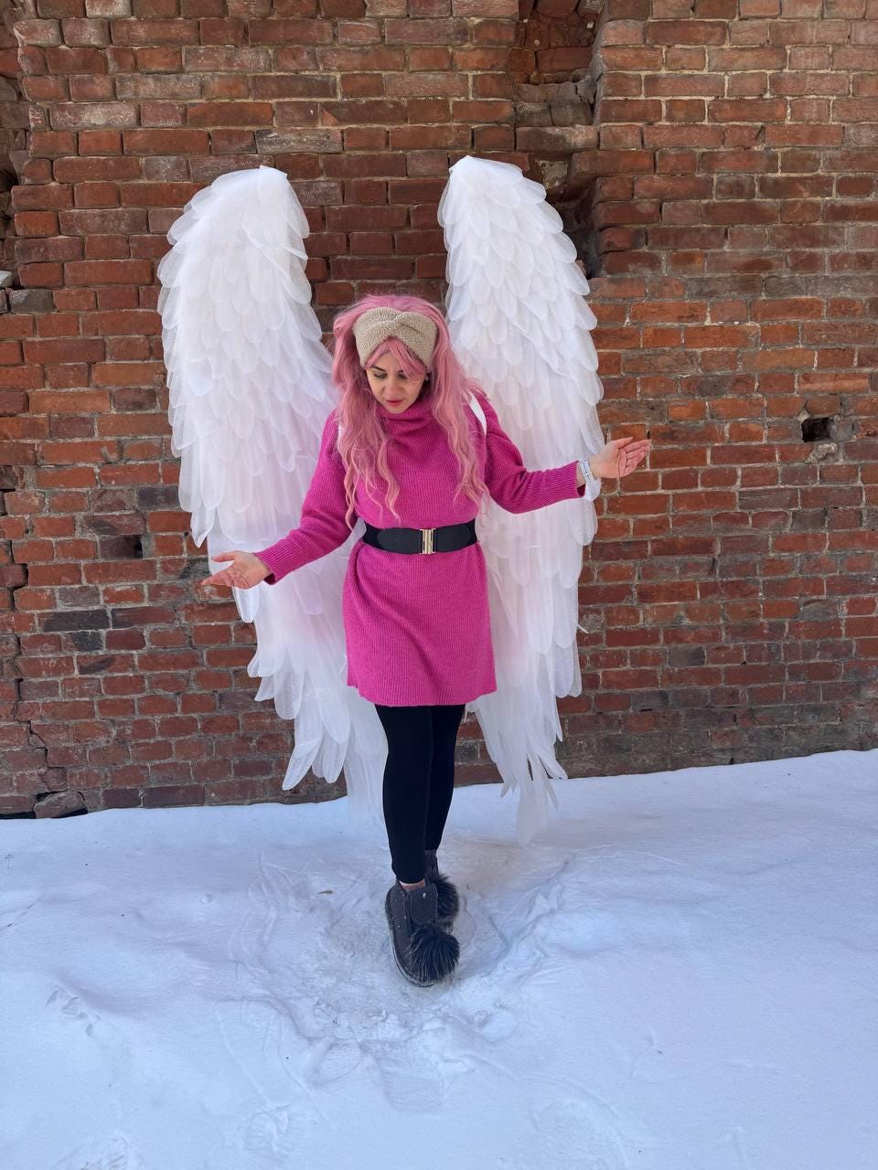 May include: A person wearing a pink dress, black leggings, and gray boots stands in front of a brick wall, with large white angel wings. The person has pink hair and a beige headband. Snow covers the ground.