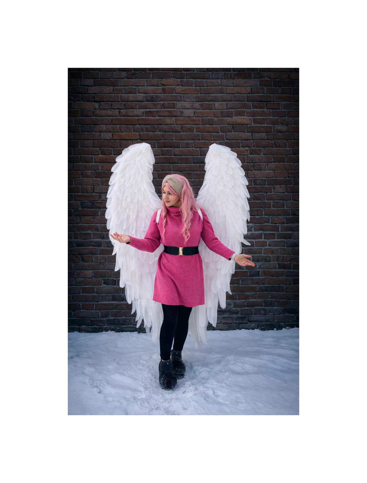 May include: A person wearing a pink dress, black leggings, and black boots, with large white feathered angel wings. The person is standing in the snow in front of a brick wall.
