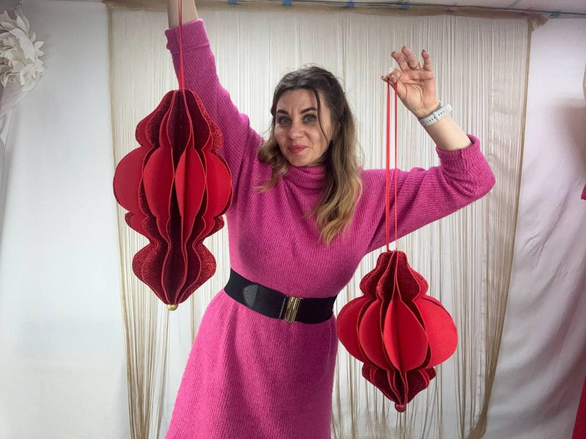 May include: Two red paper lanterns with intricate designs are suspended by red strings. The lanterns are held up by a person wearing a pink dress and a black belt. The lanterns have a layered, three-dimensional appearance.