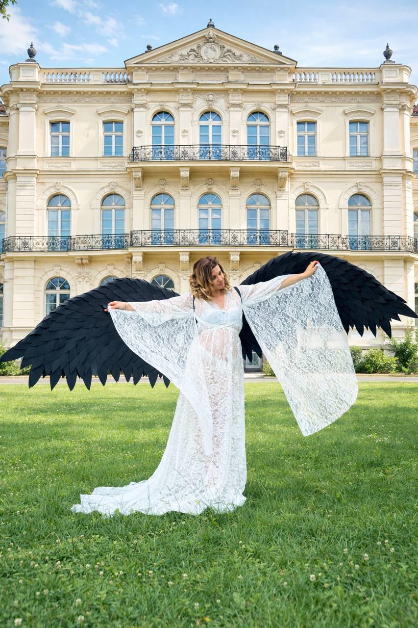 May include: A woman wearing a white lace dress and large black angel wings stands on a grassy lawn in front of a large building. The wings are detailed with a feathered design. The dress has long sleeves and a flowing skirt.