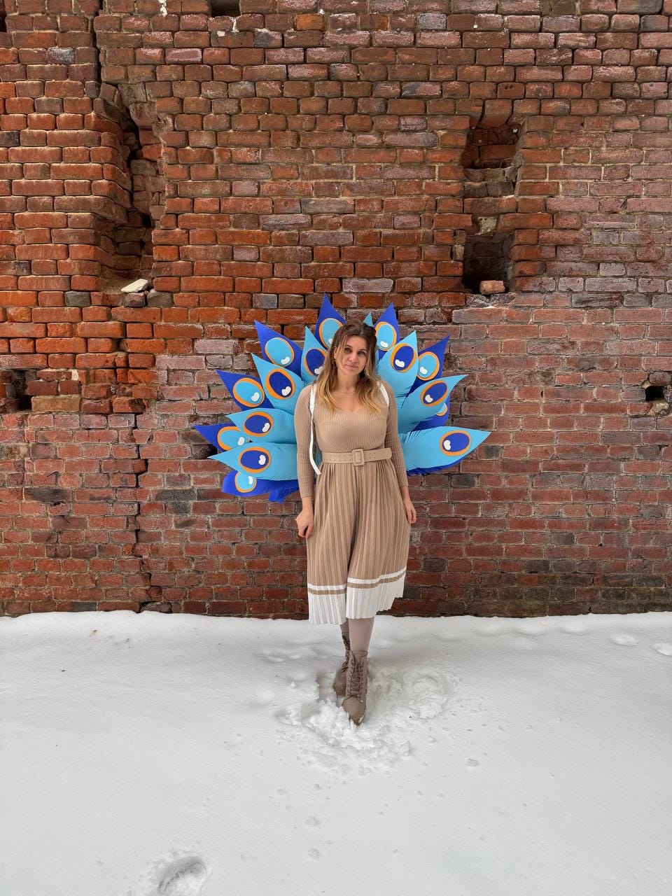 May include: A woman stands in the snow in front of a brick wall, wearing a tan outfit and a peacock-inspired costume. The costume features blue and teal feather-like shapes. The outfit includes a belted top and pleated skirt with white trim.