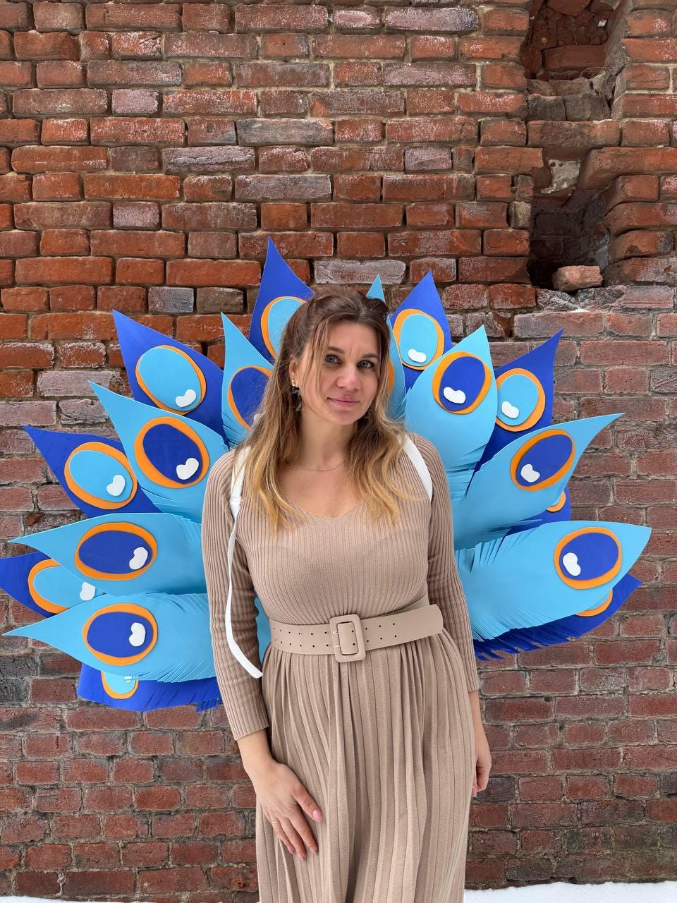 May include: A woman stands in front of a brick wall, wearing a tan dress with a matching belt. Behind her is a large, colorful peacock feather decoration in shades of blue, orange, and white. The feathers are large and stylized.