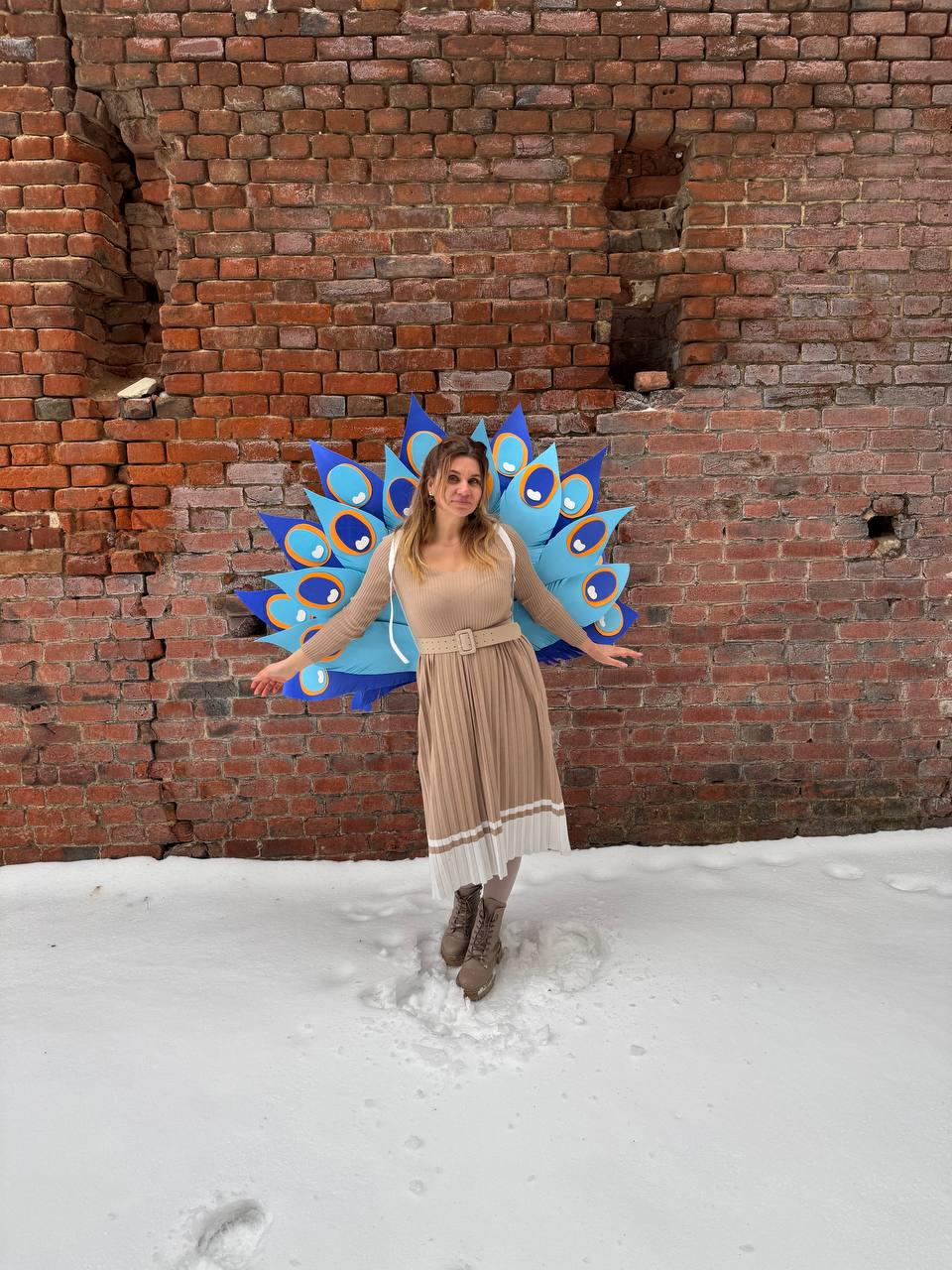 May include: A woman wearing a tan dress and brown boots stands in the snow in front of a brick wall. She is wearing a large, blue and gold peacock feather costume, with her arms outstretched.