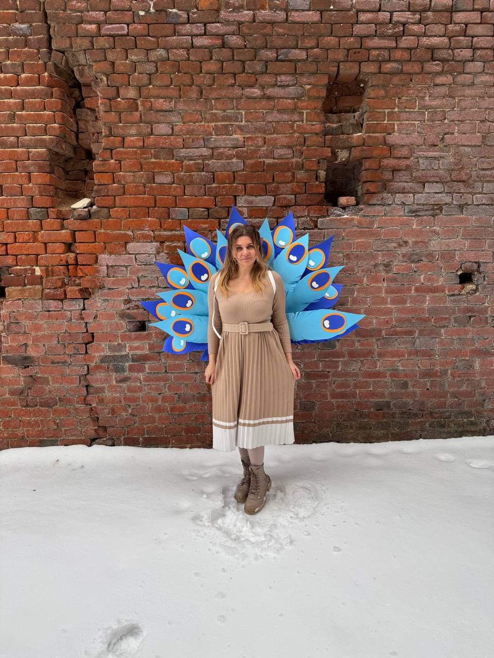 May include: A woman wearing a tan dress with a white hem and brown boots stands in front of a brick wall. She is wearing a large blue and teal peacock feather costume. The ground is covered in snow.