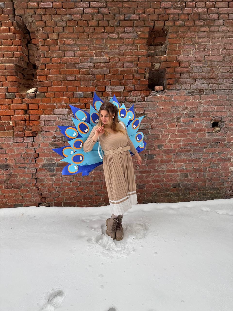 May include: A person wearing a peacock costume with a blue and gold fan-shaped design. The costume is set against a brick wall and snow-covered ground. The person is wearing a beige dress and brown boots.