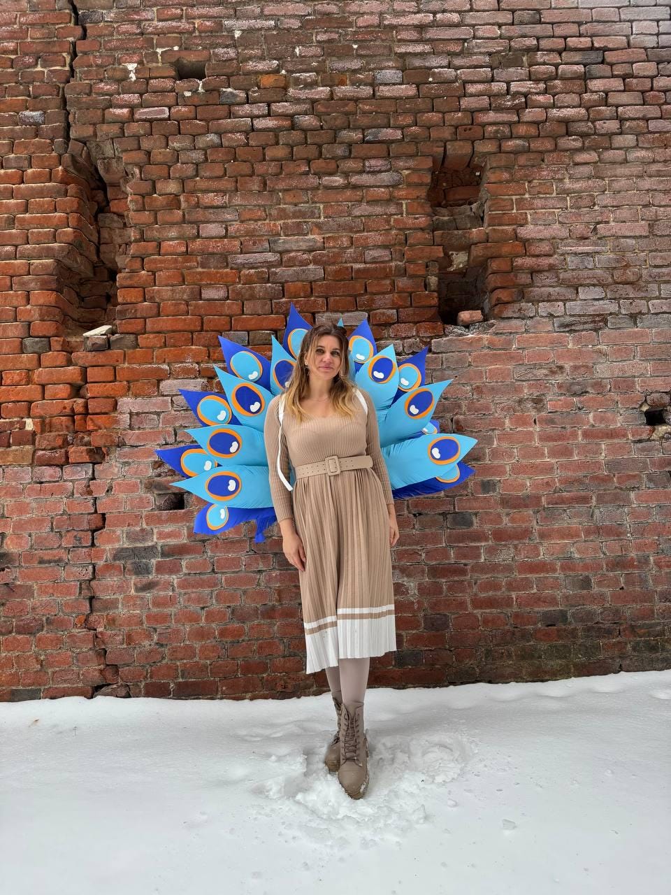 May include: A woman stands in front of a brick wall, wearing a beige dress with a belt and white accents. A large, blue peacock feather prop is behind her. The ground is covered in snow.