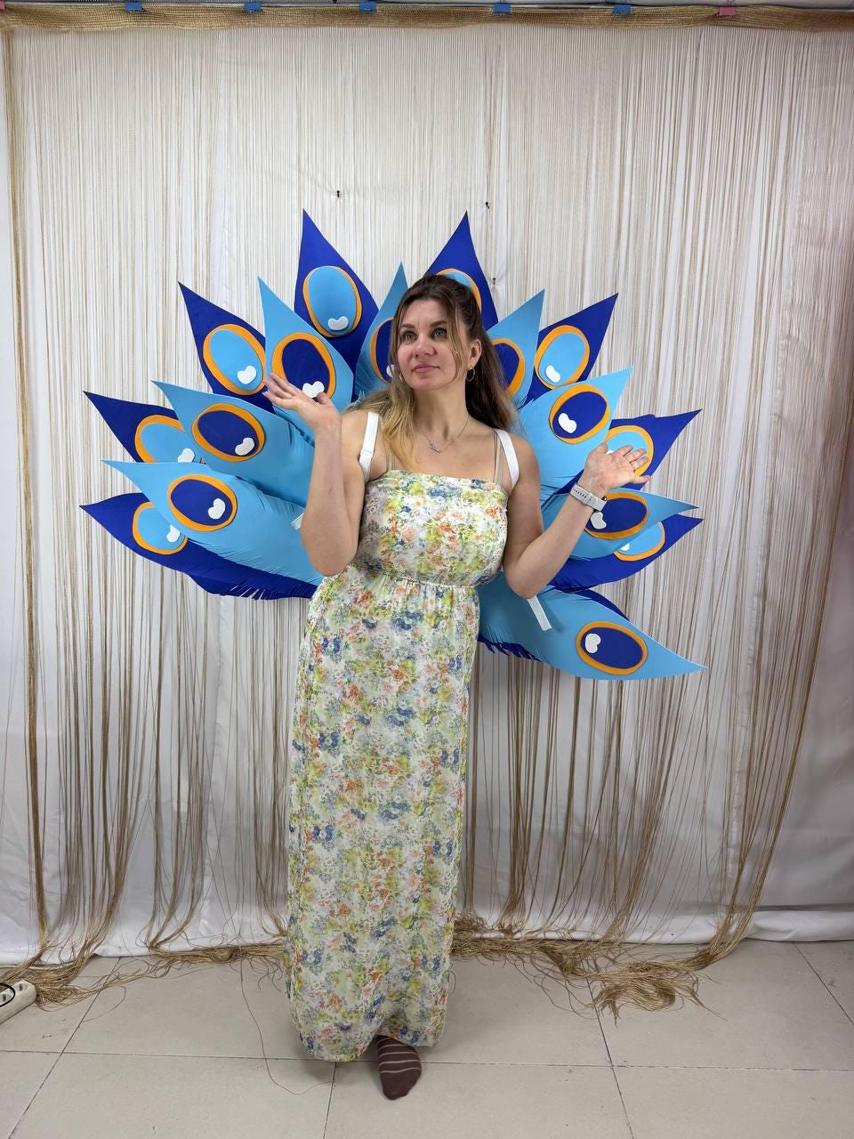 May include: A woman wearing a floral print dress and a large, blue and navy peacock feather costume. The costume has a series of large, pointed feathers with eye-like details. The background is a neutral-colored curtain.