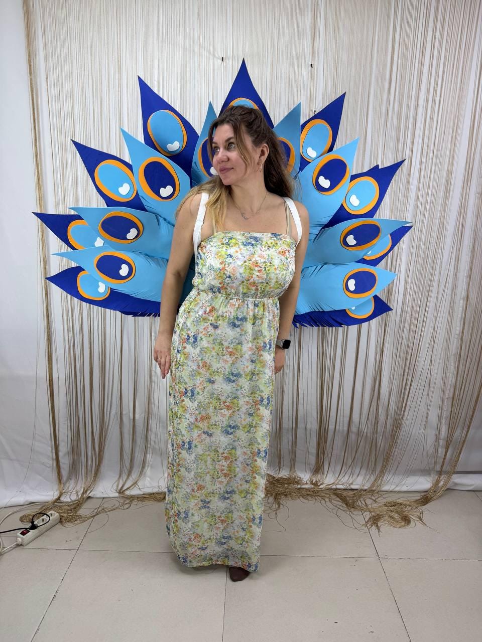 May include: A woman wearing a floral print maxi dress and a large peacock feather costume. The costume is primarily blue with orange and white accents. The dress has a white background with a floral pattern.