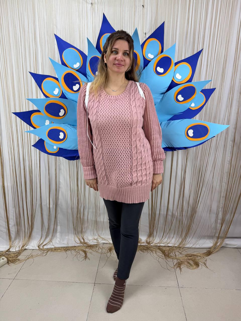May include: A woman wearing a light pink cable-knit sweater and dark gray leggings stands in front of a large peacock feather decoration. The peacock feathers are blue with orange and white accents. The background is a beige curtain.