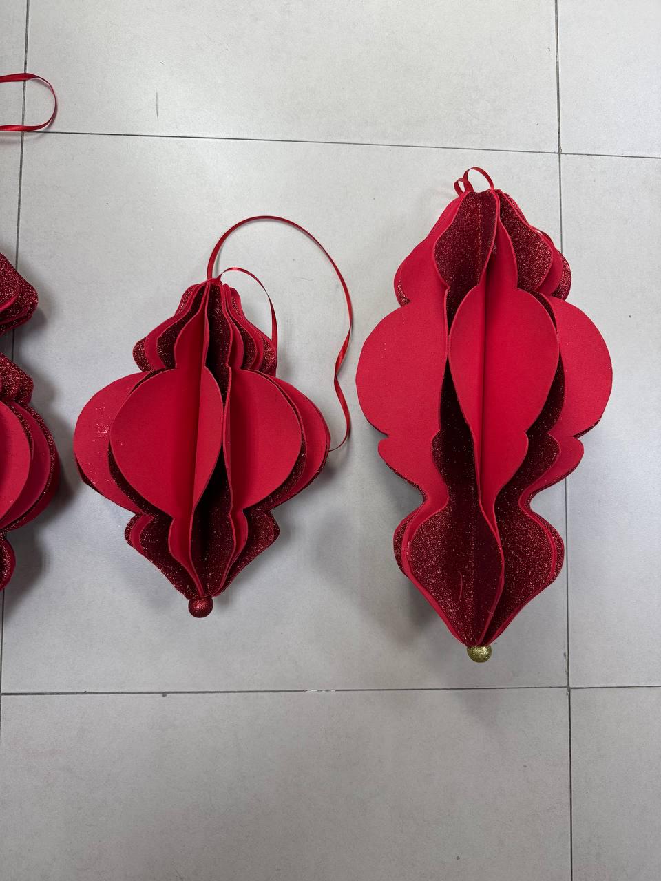 May include: Red, three-dimensional, decorative ornaments with a layered design. The ornaments have a glittery texture and are suspended by red ribbons. The ornaments are likely for holiday or seasonal decor.