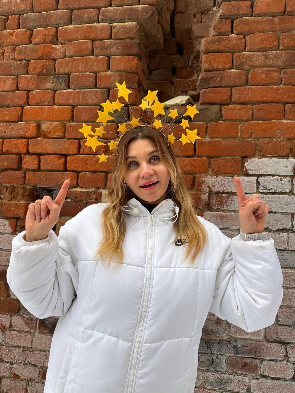 May include: A white puffer jacket with a black Nike logo on the chest. The person is wearing a yellow star-shaped headband. The background is a red brick wall.