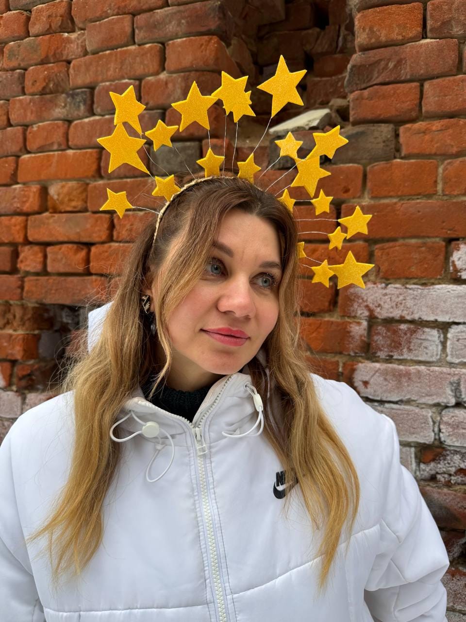 May include: A gold headband with numerous yellow star decorations. The stars are of varying sizes and are attached to the headband with thin wires. The person in the image is wearing a white jacket with a black Nike logo.
