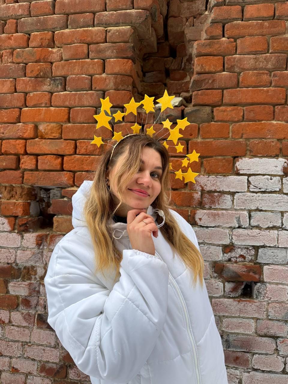 May include: A yellow starburst headband with multiple star shapes. The headband is worn by a person wearing a white puffer jacket. The background is a weathered brick wall.
