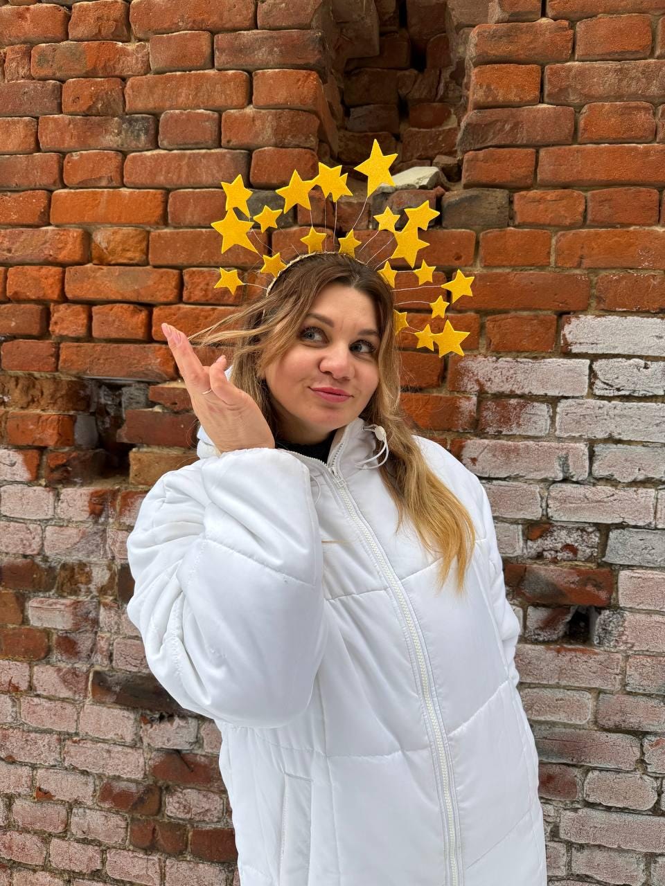 May include: A yellow starburst crown with a white headband. The crown is worn by a person wearing a white puffer jacket. The background is a weathered brick wall.