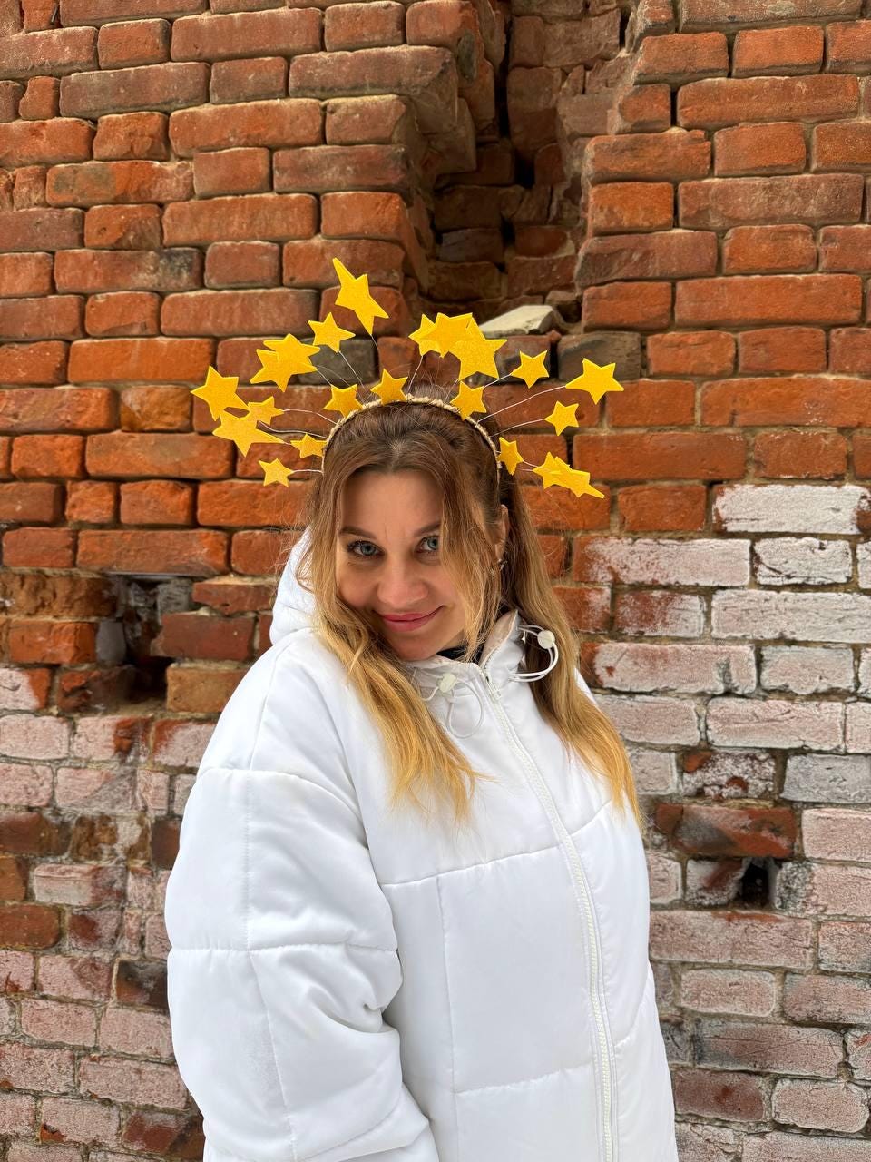 May include: A yellow starburst headband with multiple star shapes. The headband is worn by a person wearing a white puffer jacket. The background is a weathered brick wall.