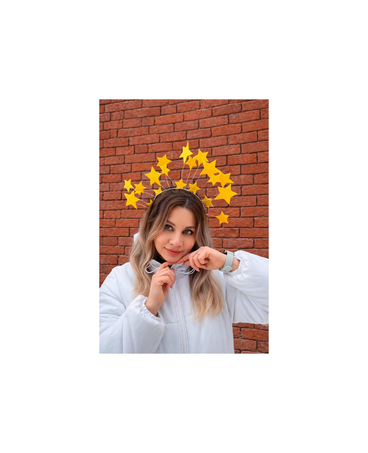 May include: A woman wearing a yellow star-shaped headband, posing in front of a brick wall. She is wearing a white jacket and a silver watch. The headband features multiple yellow stars of varying sizes.