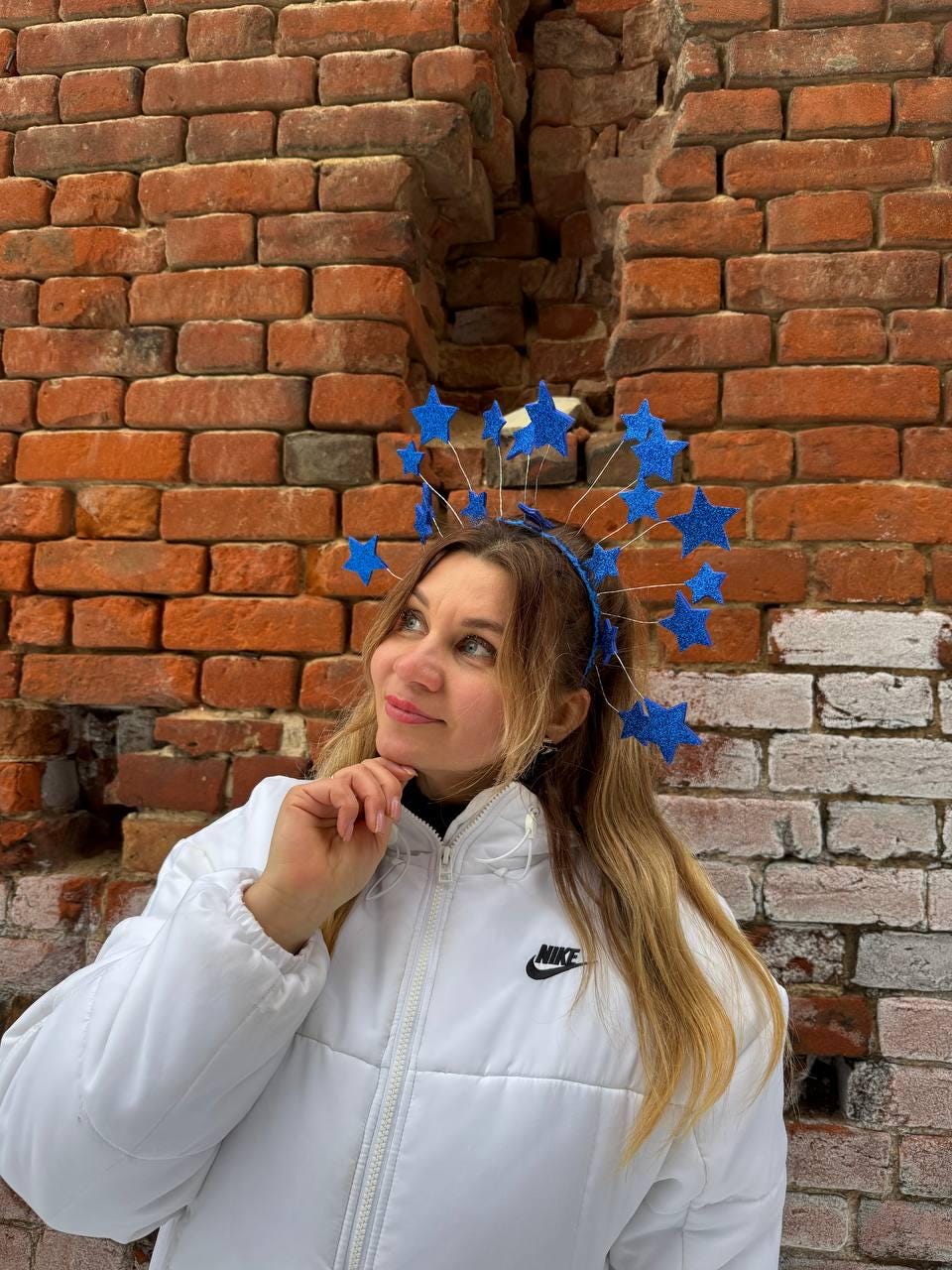 May include: A blue star headband with glittery accents. The headband is worn by a person wearing a white puffer jacket with a Nike logo. The background is a weathered brick wall.