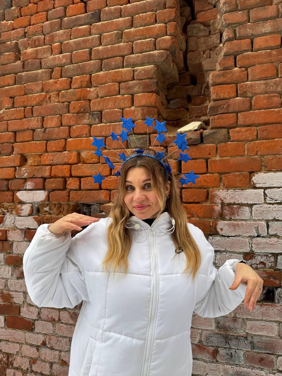 May include: A woman wearing a blue star-shaped headband and a white puffer jacket stands in front of a red brick wall. The headband features multiple blue stars attached to thin wires. The jacket has a zipper closure.