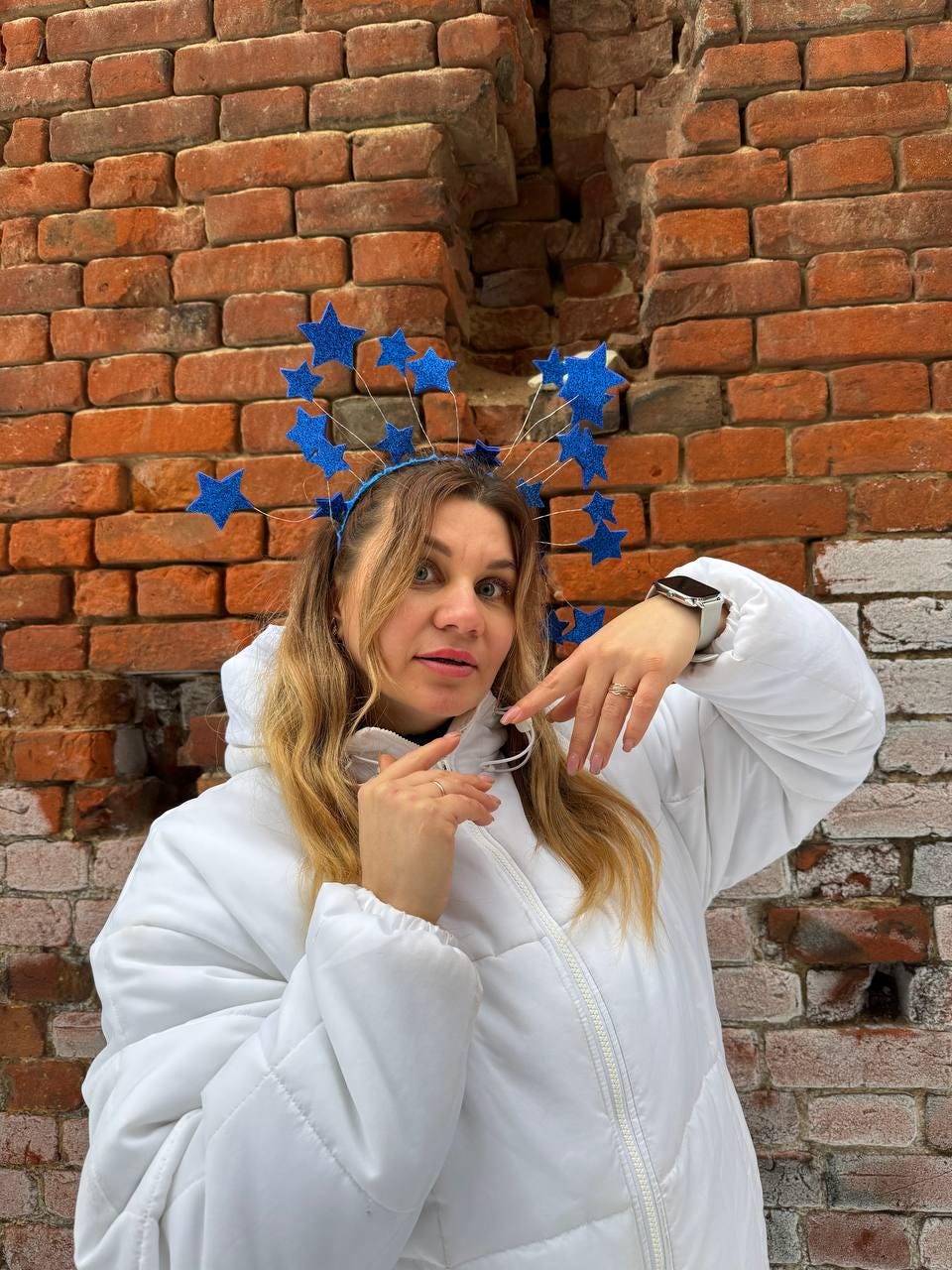 May include: A blue headband with glittery star decorations. The stars are various sizes and are attached to thin wires. The person is wearing a white hooded jacket. The background is a red brick wall.