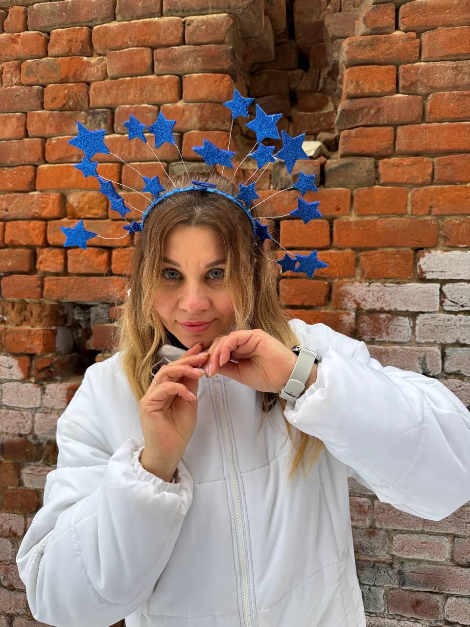 May include: A blue starburst headband with glittery stars. The headband is worn by a person wearing a white jacket. The background is a weathered brick wall.