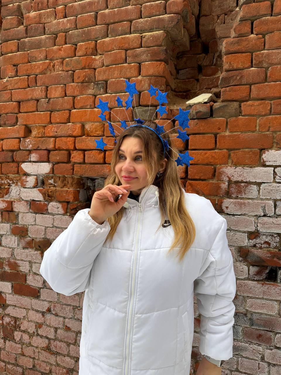 May include: A blue star-shaped headpiece with wire accents. The headpiece is worn by a person wearing a white puffer jacket. The background is a weathered brick wall.