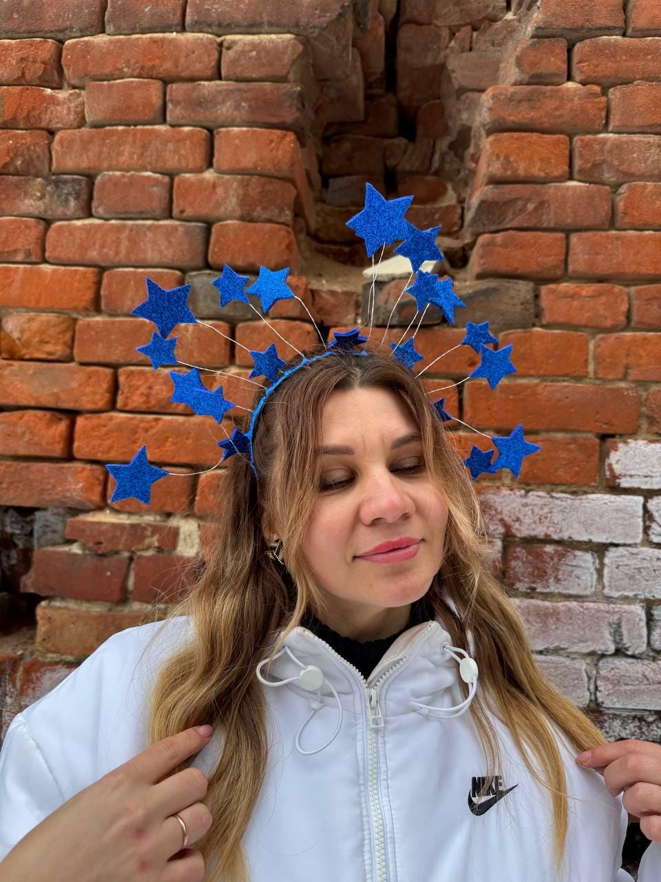 May include: A blue star headband with glitter accents. The headband features multiple blue stars attached to thin wires, creating a halo effect. The person is wearing a white jacket with a black Nike logo.