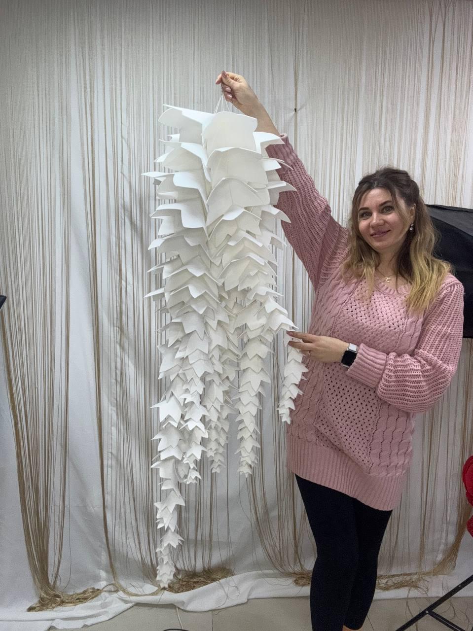 May include: A large, white, paper-crafted decorative hanging sculpture with a cascading, layered design. The sculpture is held up by a white string. The overall shape resembles a stylized, elongated plant or flower. The background is a neutral, light-colored curtain.