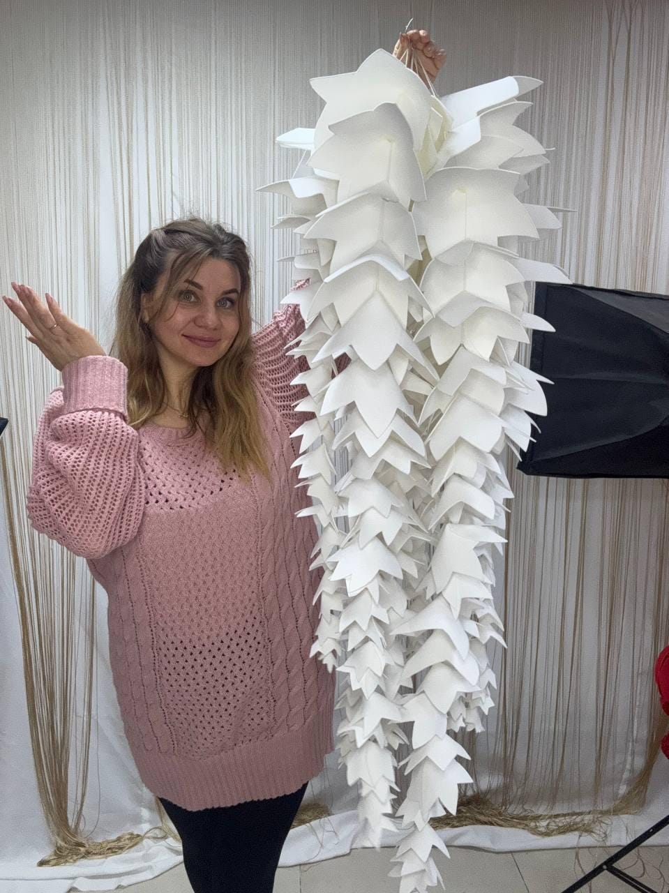 May include: A large, white paper sculpture resembling a cascading floral arrangement. The sculpture is composed of numerous layered, petal-shaped elements. The piece is held up by a person wearing a pink sweater and black pants.