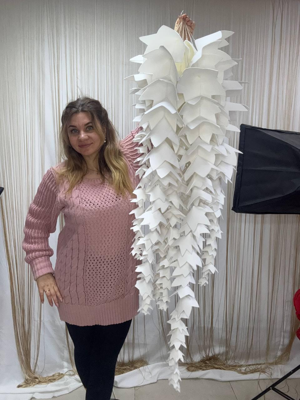 May include: A large, white paper art piece resembling a cascading floral arrangement. The piece is composed of numerous folded, leaf-like shapes, creating a layered, textured effect. The overall design is symmetrical and elegant, suitable for home decor.