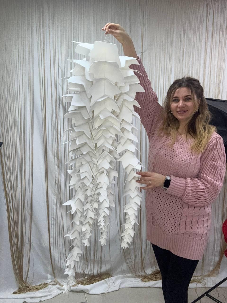 May include: A large, white, paper-crafted chandelier with a cascading, layered design. The chandelier is held up by a person. The light fixture has a unique, geometric shape, with multiple tiers of folded paper elements. The overall aesthetic is modern and artistic.