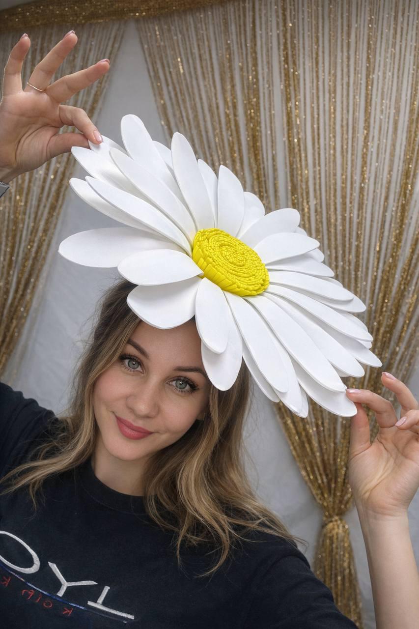 May include: A large, white daisy flower headpiece with a yellow center. The petals are layered and curved, creating a three-dimensional effect. The headpiece is worn on the head. The background is a gold curtain.