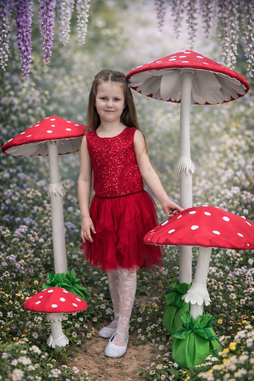 May include: A young girl in a red sequined top and tulle skirt stands among whimsical mushroom decorations. The mushrooms have red caps with white spots and white stems. The scene is set in a garden with flowers and hanging wisteria.
