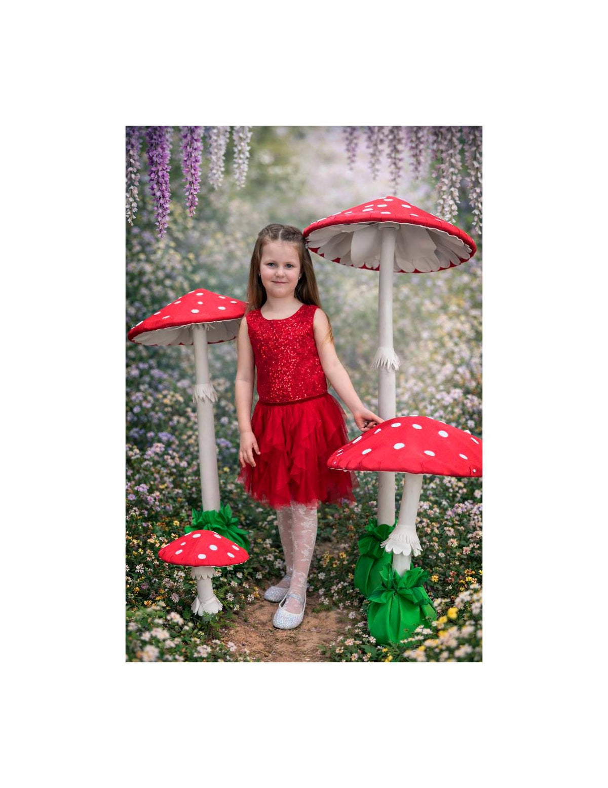 May include: A young person in a red sequined top and skirt stands among large, whimsical mushroom decorations. The mushrooms have red caps with white spots and white stems. The background features a floral setting.