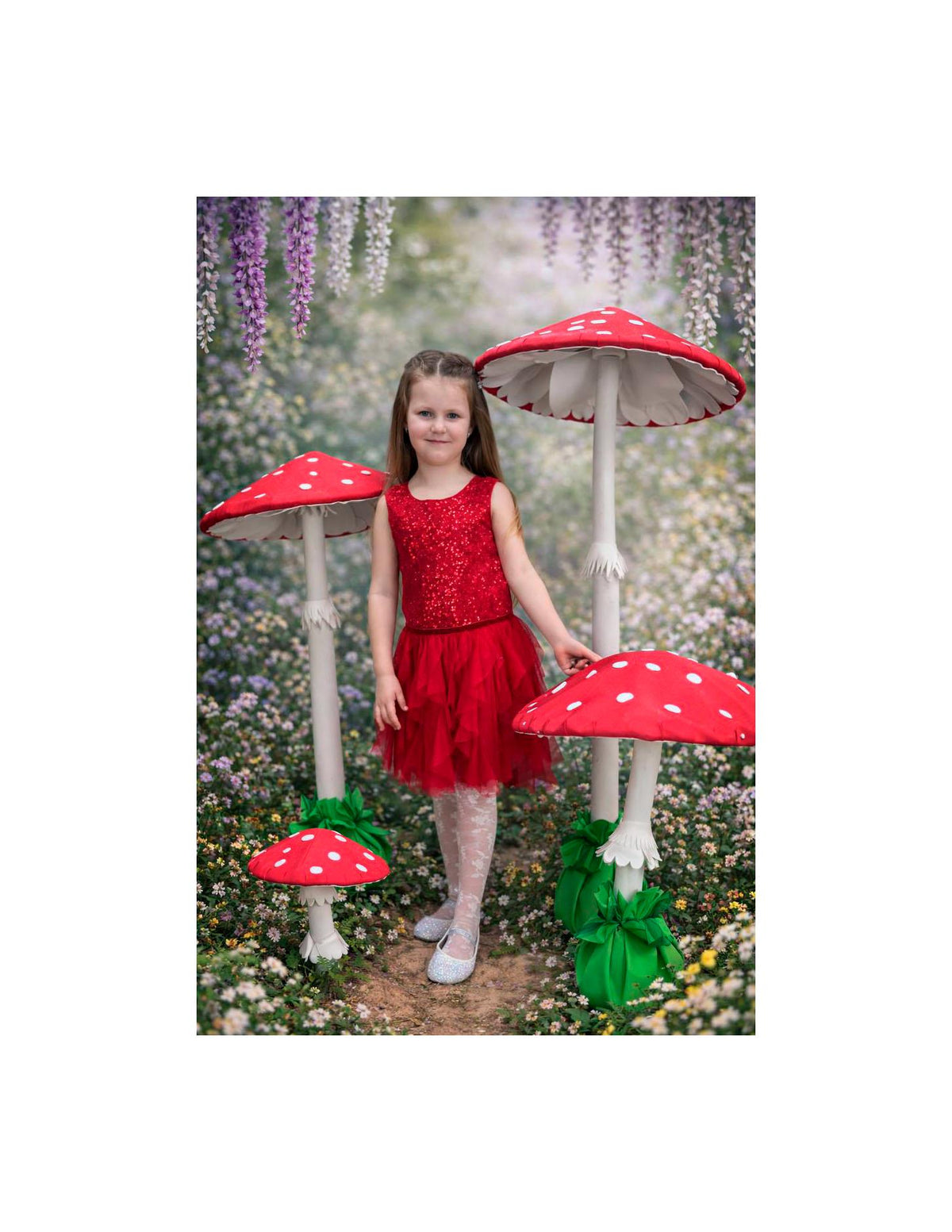 May include: A young person in a red sequined top and skirt stands among large, whimsical mushroom decorations. The mushrooms have red caps with white spots and white stems. The background features a floral setting.