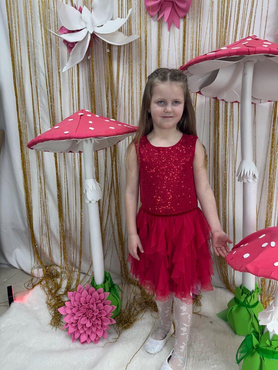 May include: A young person in a red sequined top and layered tulle skirt stands in front of whimsical mushroom decorations. The mushrooms have red caps with white spots and white stems. A large pink flower and other paper decorations complete the scene.