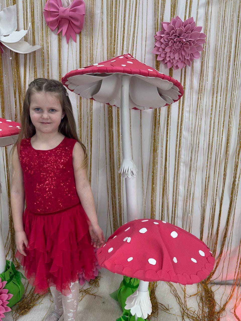 May include: A young girl in a red sequined top and tiered skirt stands near a large, whimsical mushroom prop. The mushroom has a red cap with white spots and a white stem. Decorative flowers and bows adorn the background.