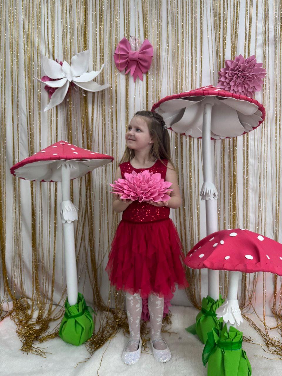May include: A young girl in a red dress stands among whimsical mushroom decorations. The mushrooms have red caps with white spots and white stems. Decorative flowers and a pink bow adorn the background, creating a fairytale theme.