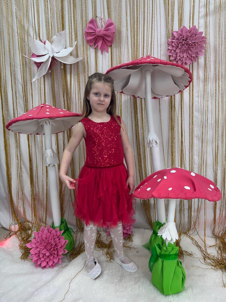 May include: A young girl in a red dress stands among whimsical mushroom decorations. The mushrooms have red caps with white spots and white stems. Decorative flowers and a bow adorn the background, creating a fairytale theme.